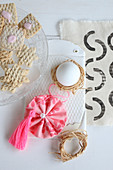 Egg in raffia nest, fabric rosette and pink tassel next to plate of biscuits