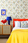 Bed with orange headboard and yellow bedspread, next to it bedside tables in the bedroom with floral wallpaper