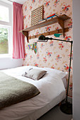 Bed below wooden shelf on wall in bedroom with floral wallpaper