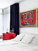 Modern art above of a queen sized bed and a red lacquered metal bedside cabinet in the bedroom