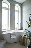 A freestanding bathtub with a freestanding tap in front of an arched window