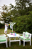 Table and seats made of white lacquered wood, with green and white cushions in the garden, in the background woman at the clothes horse