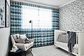 Cot against patterned wallpaper, floor-length curtains and pale grey easy chair in nursery