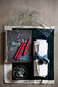 Rote Kerzen auf altem Notizbuch, Glasvase mit Blütenzweig, Weißer Stoff mit blauem Band und getrocknete Blätter