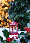 Red candle lantern on snow with holly branches in foreground