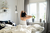 Woman sitting on rumpled bed and stretching in front of window