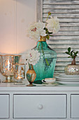 White peonies in turquoise demijohn and candle lanterns on chest of drawers