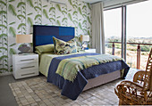 Bed with blue headboard against leaf-patterned wallpaper