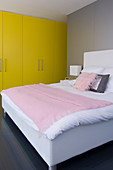 Yellow fitted wardrobes and bed upholstered in white with pink blanket and scatter cushions in modern bedroom