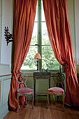 Side table and two upholstered antique chairs in front of window with long coral-red curtains