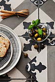 Brot, Olivengabeln und ein Schälchen Oliven auf Ornamentfliesen-Untergrund