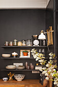 Schwarzes Regal mit Küchenutensilien und weiße Wandlampe, daneben ein Blumenarrangement
