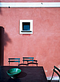 Table and chair in front of small window in pink façade