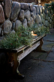 Wintry arrangement of conifer branches and candle lantern on wooden bench against stone wall