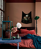 Bett vor rotbrauner Wand, im Vordergrund blauer Tisch mit Blume