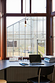 Arbeitsplatz mit Laptop und Hängeleuchte vor großen Fenstern mit Blick auf Gewächshaus
