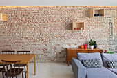 Dining-living area with wooden table and grey sofa in front of brick wall with indirect lighting