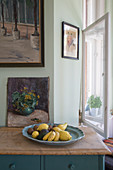 Fruit bowl and picture of flowers on top of old cabinet next to window