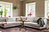 White sofa set and antique table in living room with patterned wallpaper and multiple windows