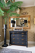 An indoor palm tree on a stela, an antique chest of drawers and a gold-framed mirror on a papered wall