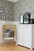 Rattan armchair next to white cupboard in corner of room with wood panelling and wallpaper