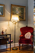 Armchair with claret-red upholstery and standard lamp below gilt-framed paintings in corner of room