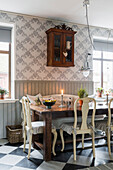 Wooden table and chairs and antique wall-mounted cabinet in kitchen with grey wainscoting and patterned wallpaper
