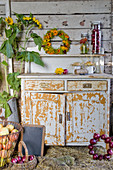 Ernte-Stilleben mit Äpfeln, roten Zwiebeln, Herbstkranz und Sonnenblumen am alten Küchenschrank