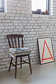 Newspapers on wooden chair and framed print against white-painted brick wall