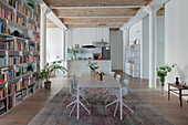 White dining table and chairs below pendant lamp in front of wall of bookshelves in open-plan interior with high ceiling
