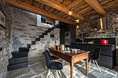Black modern kitchen cabinets, rustic wooden table and classic chairs in room with stone walls and wood-beamed ceiling