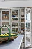 Antique crockery in display cabinet and basket of apples on kitchen island in foreground