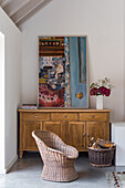 Rattan armchair and basket of firewood in front of chest of drawers with painting on top