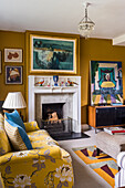 Yellow sofa, fireplace and collection of paintings on mustard-yellow wall in sitting room