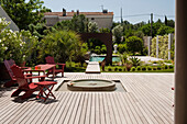 Red chairs and table around fountain on wooden terrace, garden and pool in the background
