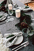 DIY garland of pine and fir branches with various conifer cones on Christmas table; place settings with pine-needle tassels as napkin rings
