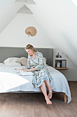 Blonde Frau sitzt mit Buch auf Bett im Dachgeschoß