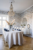 Festively set dining table, above it crystal chandelier in room with patterned wallpaper