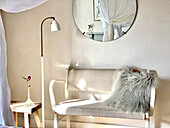 Bench with a fur blanket, floor lamp, stool with flower vase and wall mirror in the bedroom