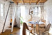 Runder Tisch mit Stühlen, Leiter zum Dachboden und Gartenzugang im Zimmer mit weiß getünchter Ziegelwand