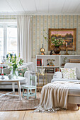 Pale upholstered furniture, bookcase and painting in living room with vintage-style wallpaper