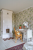 White tiled stove, dressing table and rattan chair in room with floral wallpaper