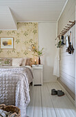 Double bed with silver-coloured bedspread and wallpaper with floral motif in the bedroom