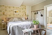 Doppelbett mit silberfarbener Tagesdecke, davor alte Holzbank mit Geranie und Tapete mit Blütenmotiv im Schlafzimmer