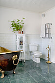 Freestanding brass bathtub, glass cabinet with houseplant on top and toilet in bathroom