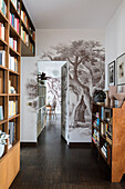 Room-high bookshelves and mural wallpaper with tree motif in hallway with open interior door