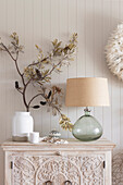 Dried banksia blossom in vase, table lamp with glass base on dresser with decoration