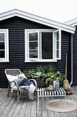 Rattanstuhl und Metallhocker auf der Terrasse vorm schwarzen Holzhaus