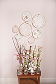 Pastel-coloured spring blossoms and twigs in bottles, above wall plate and embroidery frame with lace doily