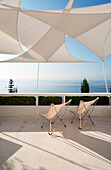 Klassikerstühle auf Terrasse mit Sonnensegeln, das Meer im Hintergrund